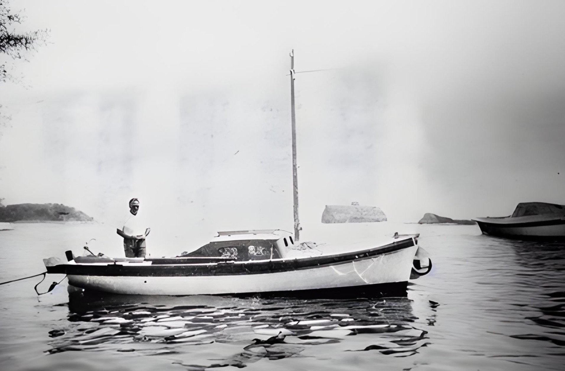 Antonio Dekovich standing at the bow of his boat, Altoal, at anchor in still water under haze.